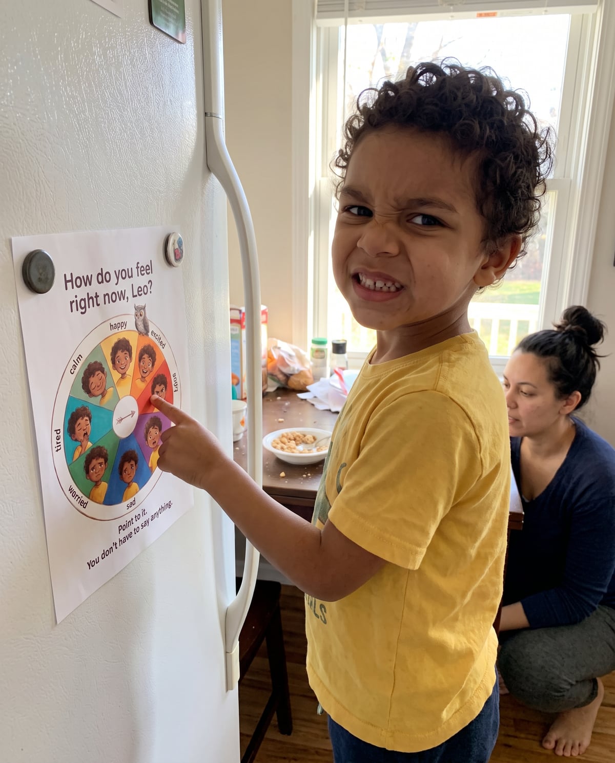 A boy at a fridge pointing at a feelings wheel printed with his own face